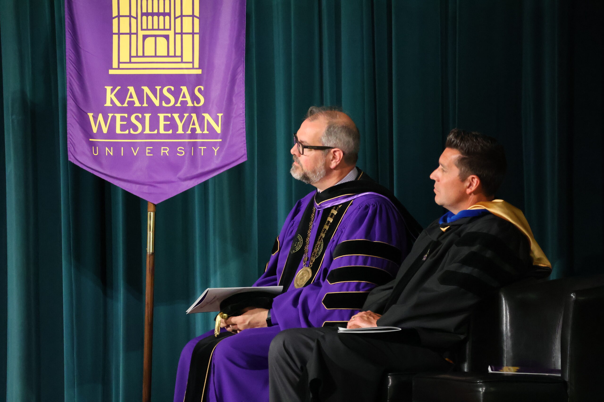Two men sitting next to university banner in grad attire